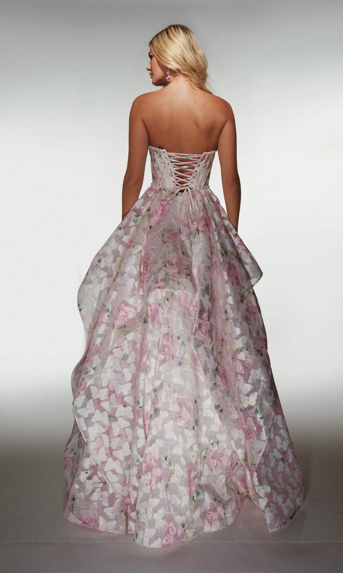 A woman stands with her back to the camera in the Alyce 62071 long prom dress, a strapless organza ball gown featuring a floral pattern and lace-up back, set against a plain background.